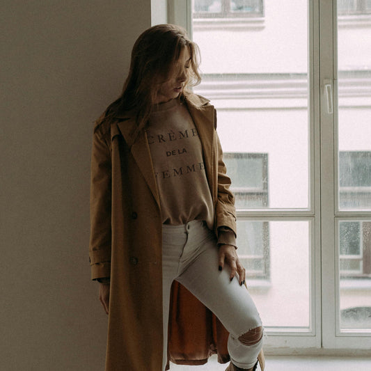 A woman wearing a brown sweatshirt with the text 'CREM DE LA CREM' on it, paired with a beige coat and white ripped jeans, standing indoors.