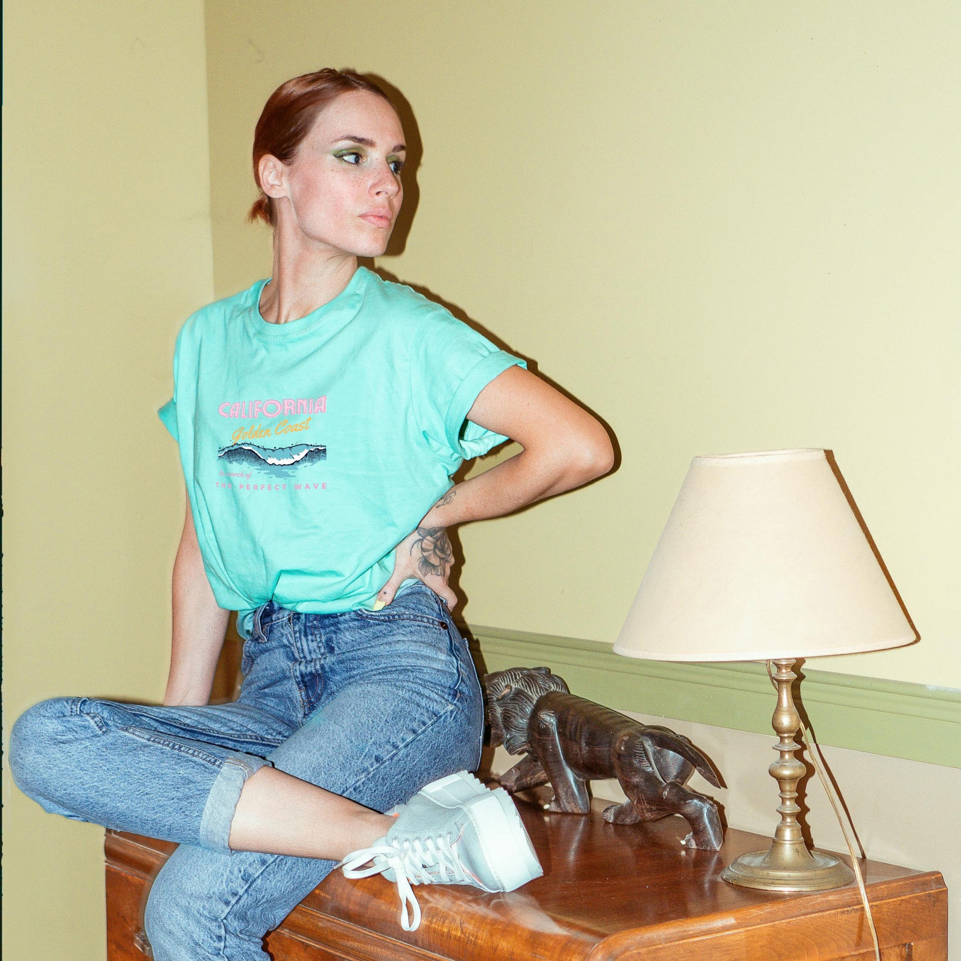 A woman sitting on a table wearing a green graphic t-shirt and blue jeans, with sneakers and a watch. She has red hair and is posing with one hand on her hip.