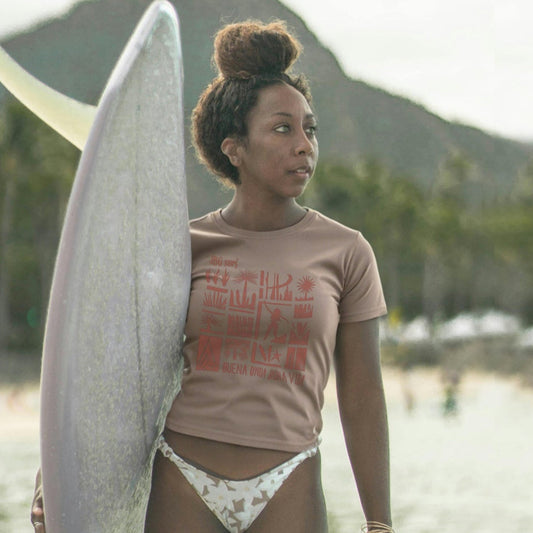 A woman standing by the beach wearing a high-waisted t-shirt with a tribal vintage-style graphic and high-waisted shorts, holding a surfboard.