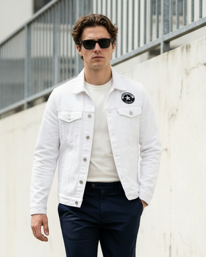 Man wearing a white denim jacket, white shirt, dark pants, and black sneakers against a white wall.