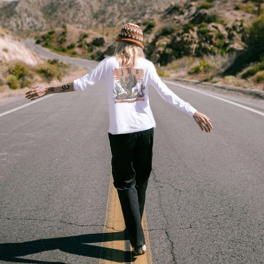 A person standing on a road wearing a white long-sleeve t-shirt with a desert-themed graphic print featuring a snake, leopard, and cactus, paired with black pants and a hat.