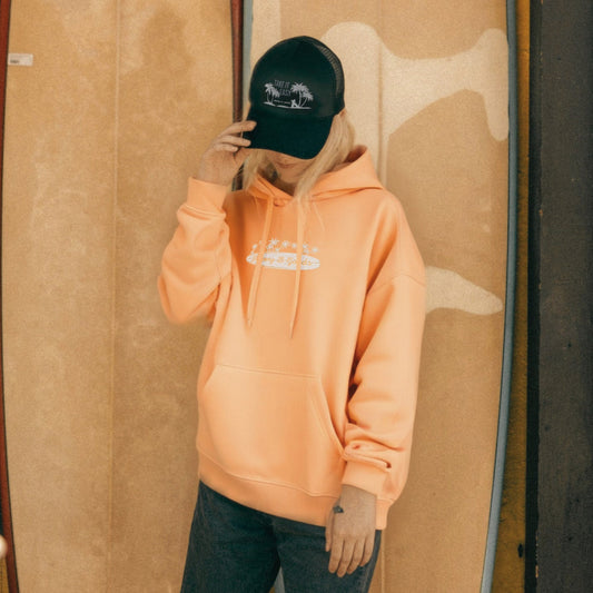 A woman wearing an orange midweight hoodie with a surf graphic print and a black cap.
