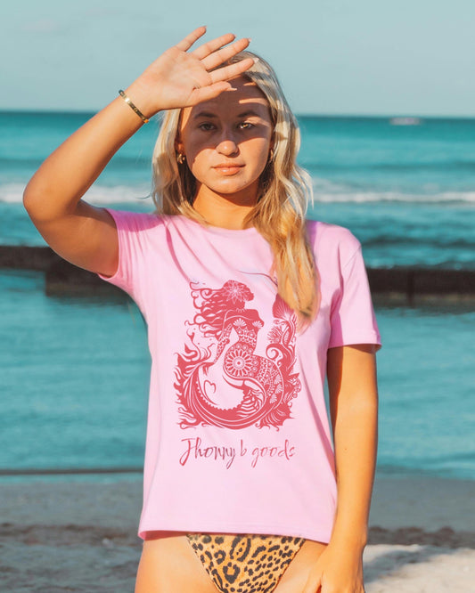 A woman standing by the beach wearing a pink oversized graphic t-shirt with a mermaid design and the text 'JBG surf & skate' on it.