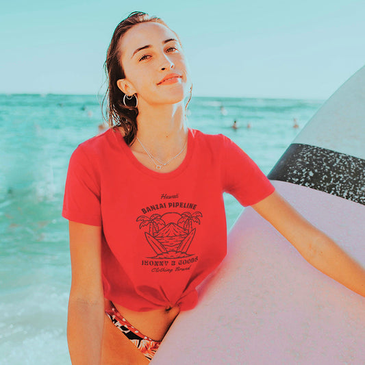 A woman standing by the sea wearing a red 'Banzai Pipeline' graphic tee with a surf design.