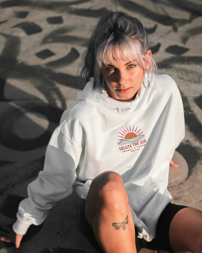 A woman sitting on a shadowed surface wearing a white hoodie with the sun and text embroidered on the back.