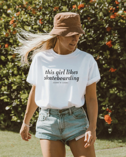 A woman wearing a white t-shirt with the text 'this girl likes skateboarding' printed on the front, paired with denim shorts and a bucket hat.