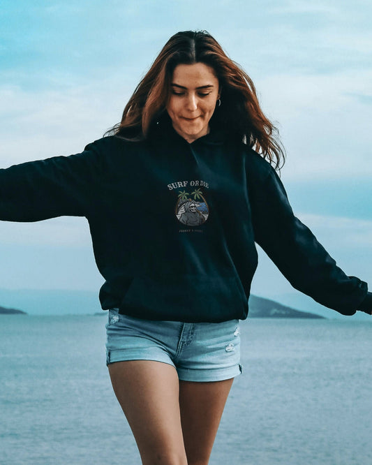 Woman in black hoodie and denim shorts standing with arms outstretched by a body of water.
